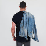 Man wearing a black t-shirt, gray jeans, and a denim jacket over his shoulder on a white background