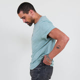 Man wearing a teal t-shirt and dark jeans standing against a white background