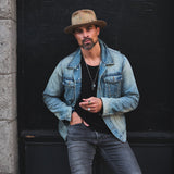 Man wearing a denim jacket and hat standing against a dark door.