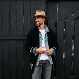 Man wearing a hat and casual clothing standing against a dark wooden wall.
