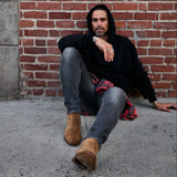 Man sitting against a brick wall wearing a black hoodie and brown boots